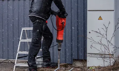 Elektricien slaat met een breekhamer een aardpen in de grond naast een woning